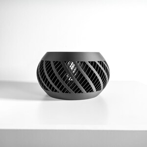 May include: A matte black spherical bowl featuring a complex, open-work lattice pattern of diagonal slats. The object has a solid, smooth rim at the top and a matching solid base, creating a hollow, cage-like structure with a dark, uniform finish.