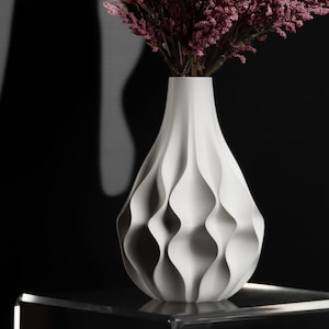 May include: A white vase with a textured, undulating vertical wave pattern on its exterior surface. The vase has a narrow neck that widens into a rounded body and is filled with a bouquet of dark pink, feathery, textured floral stems.
