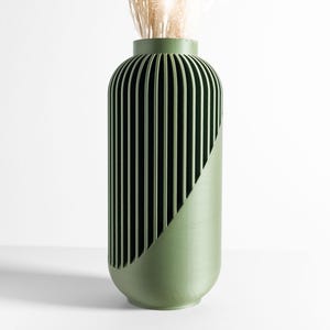May include: A cylindrical olive green vase featuring a textured exterior with vertical slats on one side and a smooth, solid surface on the other. The vase has a narrow opening at the top and contains a small amount of dried beige decorative grass.