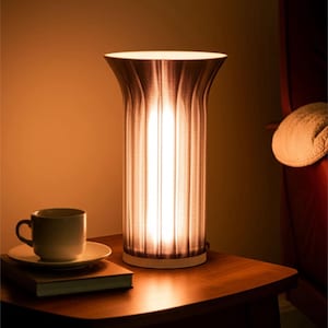 May include: A cylindrical table lamp with a textured, dark brown shade, emitting a warm glow. The lamp sits on a wooden side table, next to a cup and saucer, and a book. The lamp's height is approximately 12 inches.