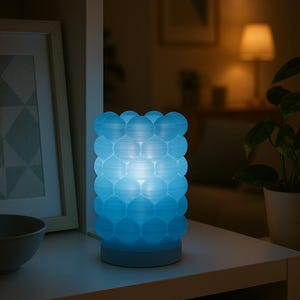 May include: A blue decorative lamp with a textured, bubble-like design. The lamp emits a soft, glowing light, creating a calming ambiance. It sits on a white shelf, with a framed picture and a potted plant in the background. The lamp is approximately 6 inches tall.