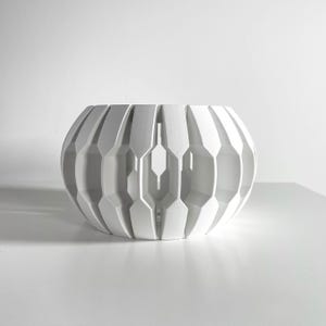 May include: A white, spherical, open-work bowl featuring a series of vertical, geometric, fin-like panels arranged in a radial pattern. The structure has a hollow center and a repeating, angular, hexagonal cutout design along its sides.