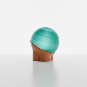 May include: A spherical object featuring a translucent, textured green glass top section with subtle horizontal banding, resting securely within a solid, smooth, matte-finished brown base that has a curved, molded shape at the bottom.