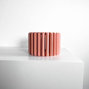 May include: A cylindrical bracelet featuring a series of vertical, faceted, salmon-pink slats arranged in a repeating pattern around the circumference, creating a structured, open-work design with a matte finish.