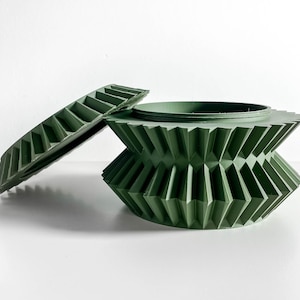 May include: A green, geometric container with a matching lid. The container has a unique, angular design with a series of triangular shapes. The lid is a flat, rectangular shape with a series of parallel ridges. The container and lid are made of a solid, matte green material.