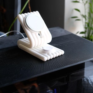 May include: A white, multi-tiered, ribbed charging stand featuring a circular magnetic wireless charger on the angled upright section and a smaller circular charging pad on the flat base, connected by a white power cable extending from the rear.