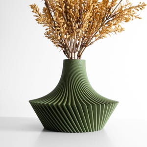 May include: A green decorative vase with a unique, geometric design. The vase has a wide, angled base and a narrower neck, with vertical lines creating a ribbed effect. The vase holds dried, yellow-brown floral stems, adding a natural element.