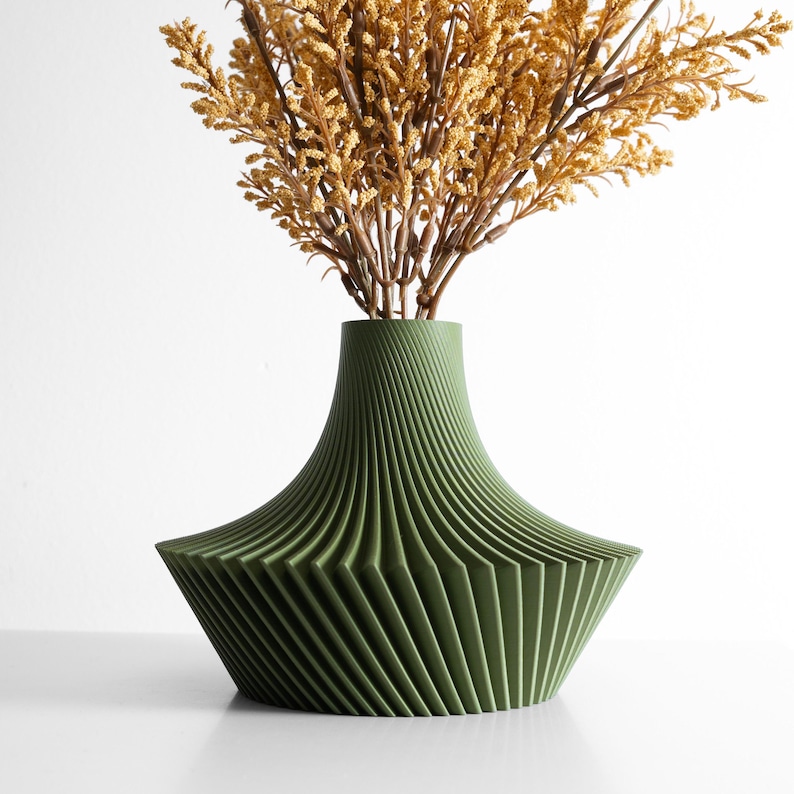 May include: A green decorative vase with a unique, geometric design. The vase has a wide, angled base and a narrower neck, with vertical lines creating a ribbed effect. The vase holds dried, yellow-brown floral stems, adding a natural element.