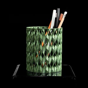 May include: A green, textured pencil holder with a geometric design. The holder contains several pencils and pens in various colors, including white, black, and brown. The holder is cylindrical and sits on a black surface.