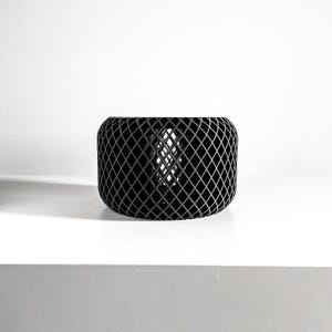 May include: A black, cylindrical decorative object with a diamond-patterned exterior. The object is made of a dark material and sits on a white surface. The design features a lattice-like structure, creating an open, airy aesthetic. The object is a modern home decor item.