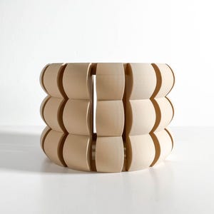May include: A cylindrical light-colored wooden object featuring a repeating pattern of vertical slats with rounded, convex outer surfaces. The structure is composed of three stacked horizontal rows of these curved wooden segments, creating a hollow, open-ended form with a smooth, matte finish.