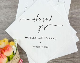 May include: A stack of white paper napkins featuring the black printed text "she said yes" in a cursive script, followed by the names "HAISLEY and HOLLAND", a small heart icon, and the date "MARCH 17, 2026" in a clean sans-serif font.