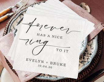 May include: A stack of white paper napkins with the text "forever has a nice ring to it" in elegant black script. The napkins are on a decorative plate with a floral design, set for a special occasion.