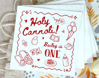 May include: White paper napkins with a red design. The napkins feature the text "Holy Cannoli!" and "Bailey is ONE", along with illustrations of baby items and food. The napkins are stacked.