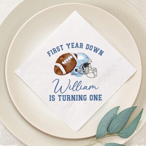May include: A white square napkin with a football and helmet design. The text reads "FIRST YEAR DOWN" and "William IS TURNING ONE" in blue. The napkin is on a white plate with green leaves.