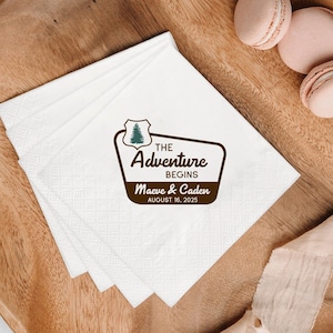 May include: White paper napkins with a brown and green graphic that reads "The Adventure Begins" with the names Maeve & Caden and the date August 16, 2025. The napkins are stacked on a wooden surface.