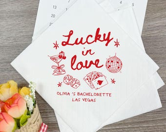 Op de afbeelding: Witte papieren servetten met rode tekst en illustraties. De zin "Lucky in Love" is geschreven in een decoratief lettertype, samen met een cocktail, dobbelstenen, speelkaarten en een discobal. De tekst "Olivia's Bachelorette Las Vegas" is ook opgenomen.