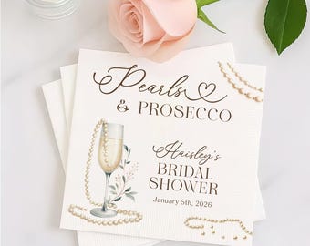May include: White paper napkins with the text "Pearls & Prosecco" and "Haisley's Bridal Shower" with a champagne glass illustration and pearl accents. A pink rose and a candle are in the background.