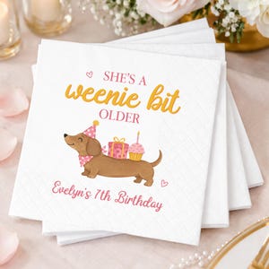 Könnte beinhalten: Ein Stapel weißer, quadratischer Papierservietten mit einer Illustration eines braunen Dackels, der einen rosa gepunkteten Partyhut und ein Halstuch trägt. Der Text lautet SHE'S A weenie bit OLDER und Evelyn's 7th Birthday, mit Geschenk und Cupcake.