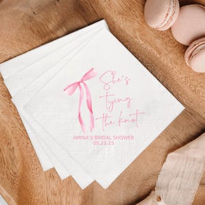 May include: White paper napkins with a pink watercolor bow and the text "She's Tying the Knot" in a script font. The napkins are for Amina's bridal shower on 09.23.25. Pink macarons are in the background.
