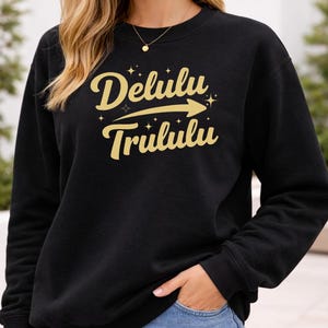 May include: Black crew neck sweatshirt with the text "Delulu Trululu" in a gold script font with an arrow. The sweatshirt is a solid black color. The text is surrounded by small starbursts.