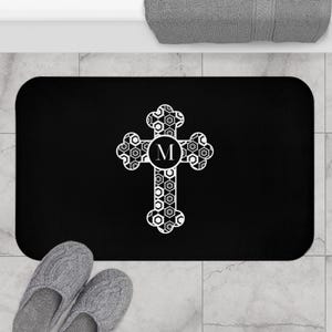 Puede incluir: Alfombra de baño rectangular negra con un diseño de cruz blanca. La cruz tiene un centro circular con la letra "M" y un patrón geométrico. Se ven zapatillas grises en la parte inferior.