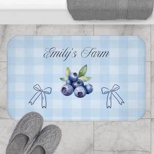 以下が含まれることがあります： 薄い青色のギンガムチェック柄の長方形バスマット。 マットには、ブルーベリーの水彩イラストと「Emily's Farm」の文字が描かれています。 ブルーベリーのイラストの両側には、2つのリボンのイラストがあります。