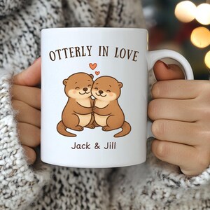 May include: White ceramic mug featuring a cartoon illustration of two hugging otters with the text "Otterly in Love" and the names "Jack & Jill". The otters are brown with pink accents, and a small heart floats above them.