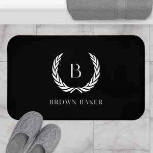 May include: Black rectangular bath mat with rounded corners. The mat features a white laurel wreath design with the letter "B" in the center, and the text "BROWN BAKER" below. Gray slippers are in the foreground.