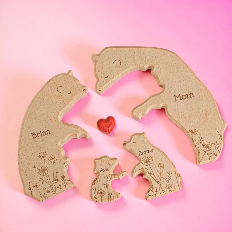 Puede incluir: Figuras de madera de una familia de osos con detalles florales y nombres grabados: Brian, Mam&aacute;, Emma y Ava. Un peque&ntilde;o coraz&oacute;n rojo se coloca entre las figuras. Los osos est&aacute;n sobre un fondo rosa.