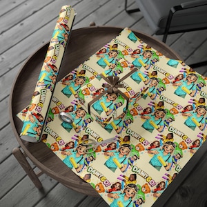 Peut inclure: Un rouleau de papier cadeau et une boîte cadeau emballée avec le même papier, présentant un motif répétitif de dessin animé avec une personne en chemise bleue tenant un tube jaune, un ours brun et le texte "DAMY GO!" sur fond crème.
