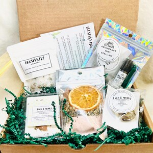 May include: A brown cardboard box filled with various items for spiritual rituals. The box contains packets of ritual salt, spell kits, dried orange slices, and green paper filler. The items are labeled with text such as "Prosperity" and "Luck & Money".