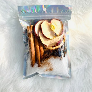 May include: A clear, sealed bag filled with dried apple slices, cinnamon sticks, and white granules. The bag has a holographic, iridescent sheen. The contents are arranged to showcase the natural colors and textures of the ingredients.