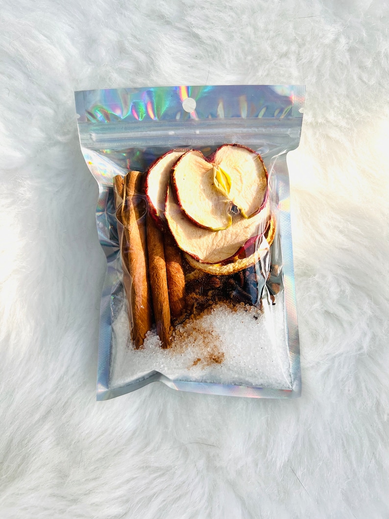 May include: A clear plastic bag filled with ingredients for a spiced apple cider drink. The bag contains dried apple slices, cinnamon sticks, star anise, cloves, and sugar.