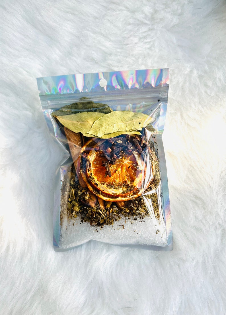 May include: A clear plastic bag filled with a variety of dried ingredients, including orange slices, cinnamon sticks, bay leaves, and cloves. The bag is sitting on a white fluffy surface.