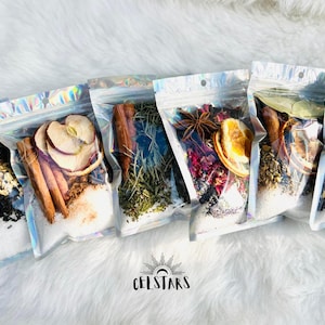 Könnte beinhalten: Fünf transparente Plastiktüten gefüllt mit verschiedenen getrockneten Zutaten, darunter Zimtstangen, Trockenfrüchte und Kräuter. Die Tüten sind in einer Reihe auf einer weißen, flauschigen Oberfläche angeordnet.