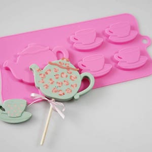 May include: Pink silicone mold with four teacup shapes and one teapot shape. A green and white teapot lollipop is on a stick.