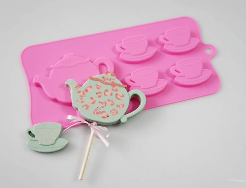 May include: Pink silicone mold with four teacup shapes and one teapot shape. A green and white teapot lollipop is on a stick.