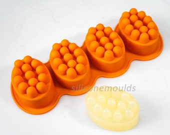 Puede incluir: Molde de silicona naranja con cuatro cavidades ovaladas, cada una con protuberancias elevadas. El molde está diseñado para hacer barras de jabón de masaje. Una barra de jabón blanca con protuberancias elevadas se muestra junto al molde.
