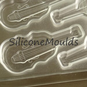 May include: Clear silicone mold with four guitar-shaped cavities. The mold is designed for making chocolate or other edible treats. The text "Silicone Moulds" is printed on the mold.