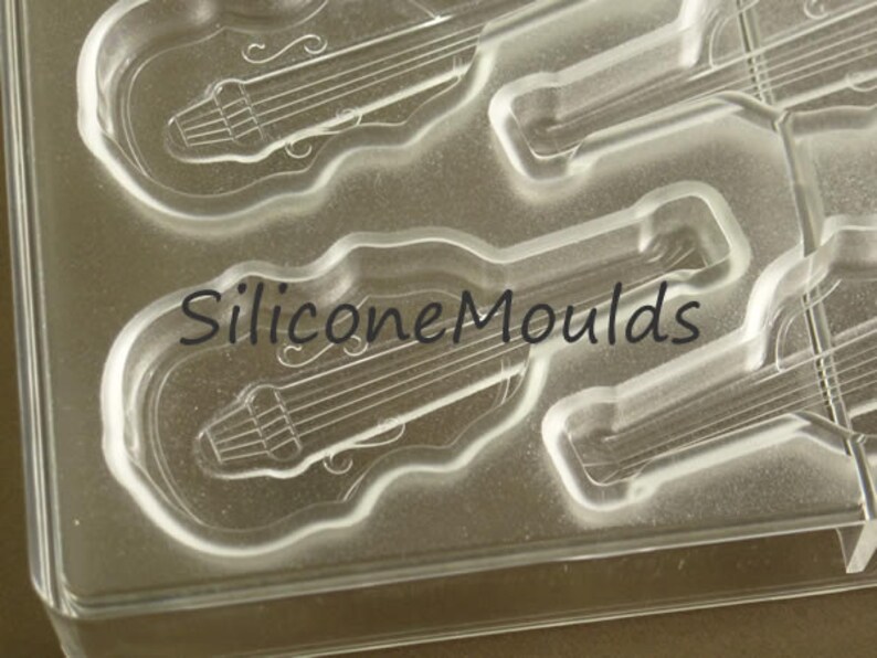 May include: Clear silicone mold with four guitar-shaped cavities. The mold is designed for making chocolate or other edible treats. The text "Silicone Moulds" is printed on the mold.