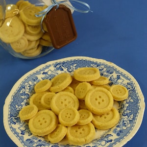 May include: A blue and white patterned plate with a pile of round, yellow button-shaped biscuits. The biscuits are arranged in a random pattern on the plate.