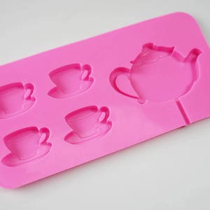 May include: Pink silicone mold with five cavities shaped like teacups and a teapot. The mold is designed for making chocolate or candy.