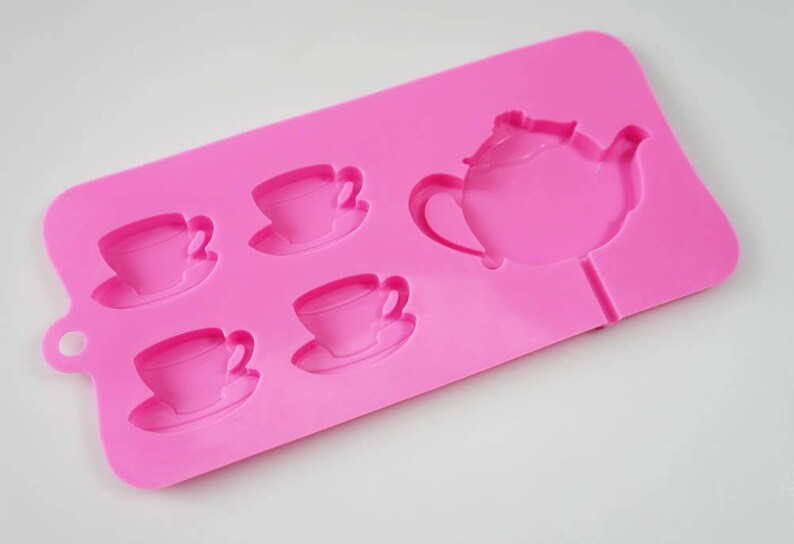 May include: Pink silicone mold with five cavities shaped like teacups and a teapot. The mold is designed for making chocolate or candy.
