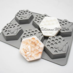May include: A grey silicone mould with six hexagonal cavities, each featuring a honeycomb pattern and a bee design. The mould holds several soaps, some white and some with a honey-like colour and glitter. The text "Silicone Molds" is visible.
