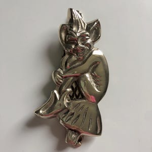 May include: A silver-coloured metal figurine of a seated imp or goblin. The imp has pointed ears, a mischievous grin, and is wearing a pointed hat. The figurine is detailed with textured surfaces and is likely a decorative item.