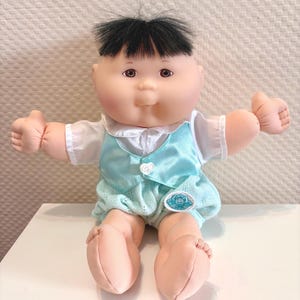 May include: A Cabbage Patch Kids doll with black hair, brown eyes, and a round face. The doll wears a white short-sleeved shirt under a light blue satin vest and matching light blue knit shorts with a small circular logo on the left leg.
