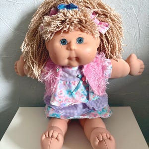 May include: A doll with blonde yarn hair, blue eyes, and a soft fabric body. The doll wears a pink fuzzy vest over a dress featuring a purple gingham bodice and a floral skirt with blue, pink, and green patterns. A small blue bow is in the hair.