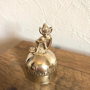 May include: A gold-coloured trinket box with a seated fairy figurine on top. The box has a rounded shape with etched detailing around the base. The fairy has detailed wings and is in a seated position.