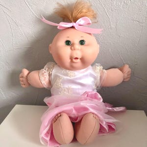 May include: A vintage doll with a round face, green eyes, and a small open mouth. The doll has light brown hair tied with a pink ribbon. It wears a white dress with lace sleeves and a pink satin skirt. The doll is sitting.